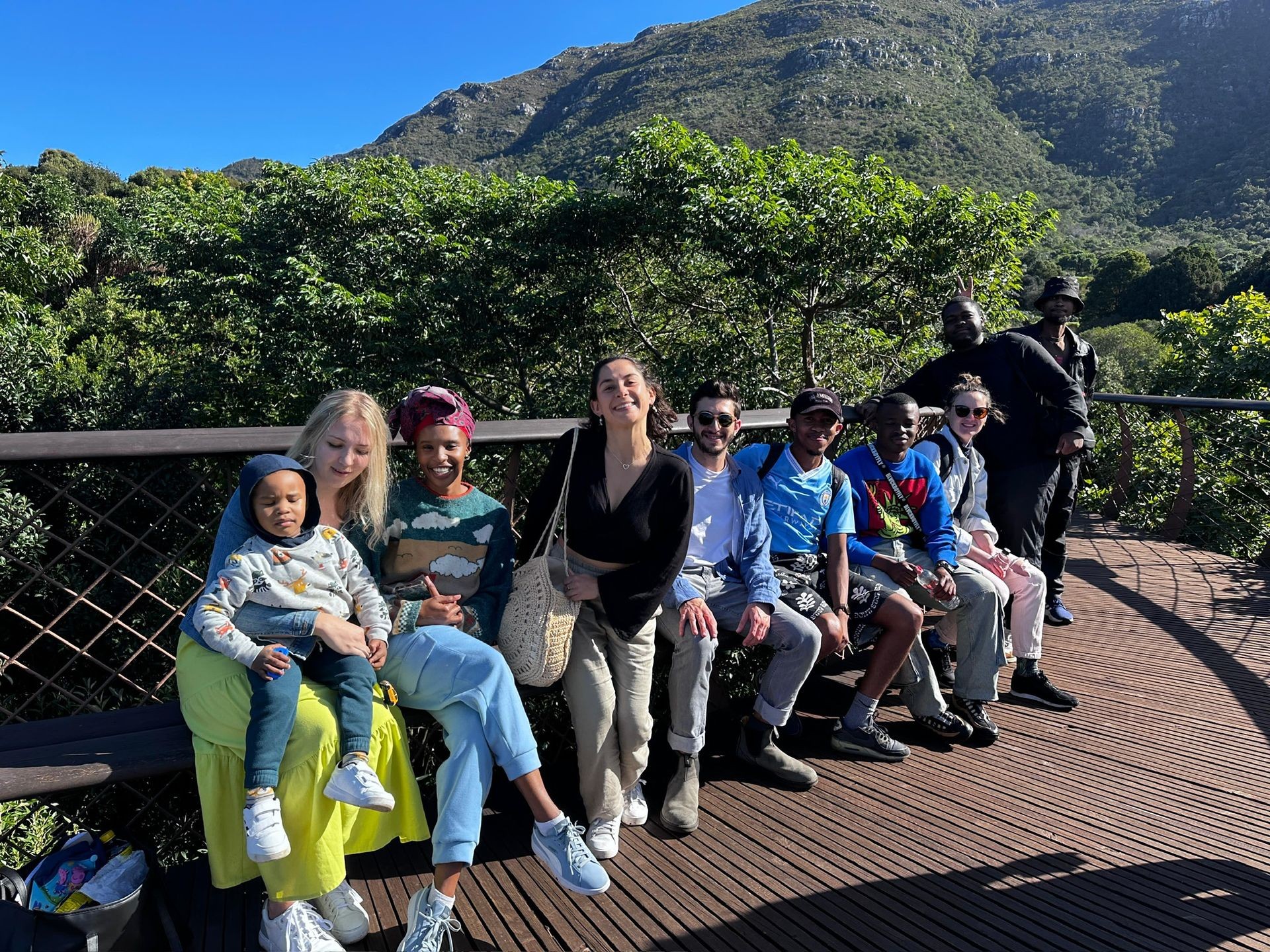 Group of people sitting on a wooden walkway with mountains and greenery in the background.