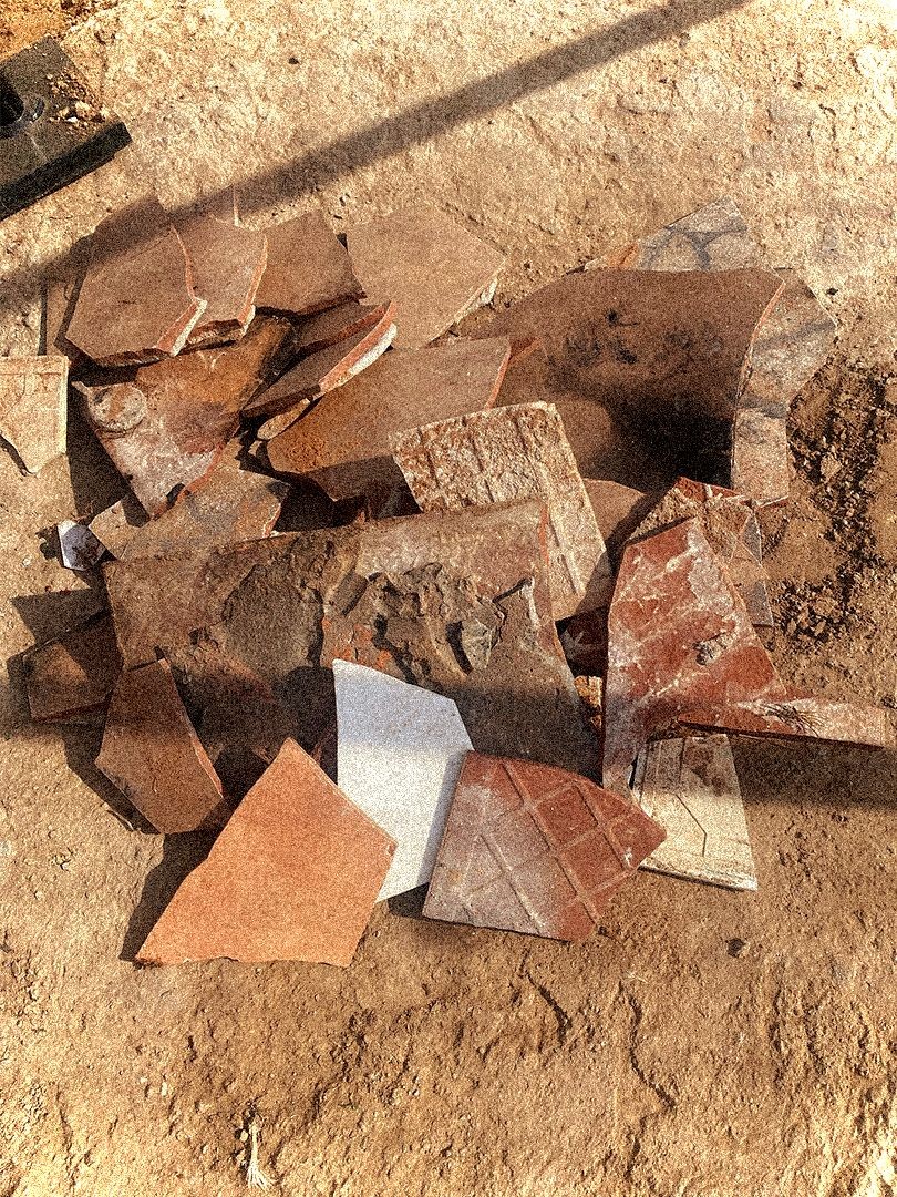 Pile of broken ceramic tiles on a dirt surface with sunlight casting shadows over the area.