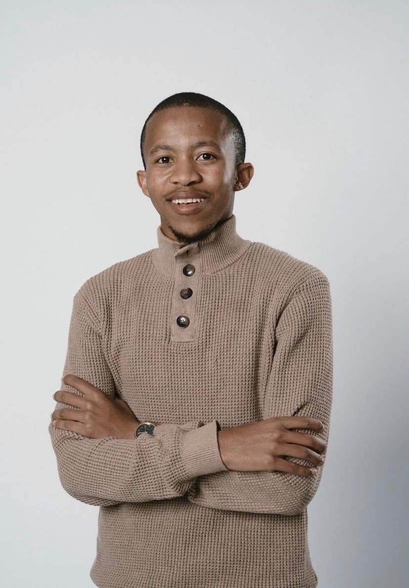 Man in a beige button-up sweater standing with arms crossed and smiling against a plain background.